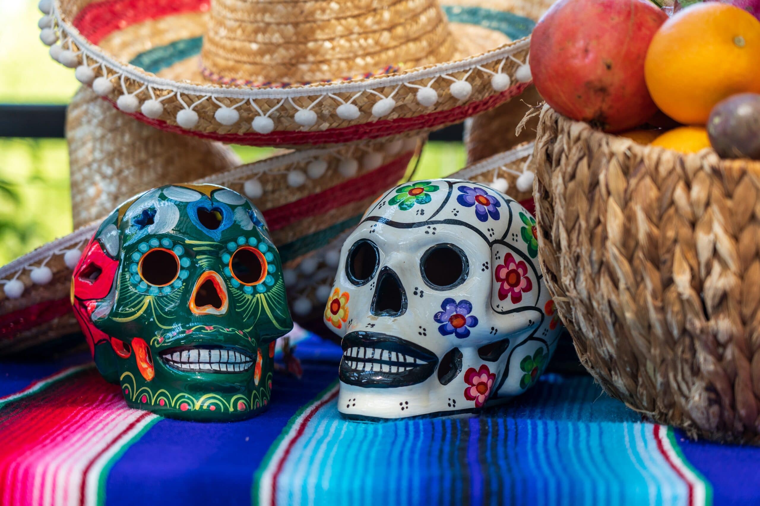 Colorful Día de los Muertos skulls with sombrero, fruit basket, and textile.