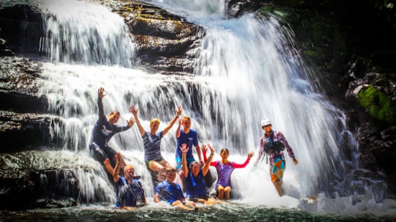 Group of people enjoying a waterfall pool, sitting and standing beneath cascading water.