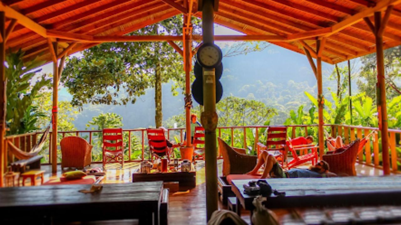 Open-air eco-lodge deck with wooden furniture, hammocks, and lounge chairs overlooking a lush tropical rainforest and mountains.